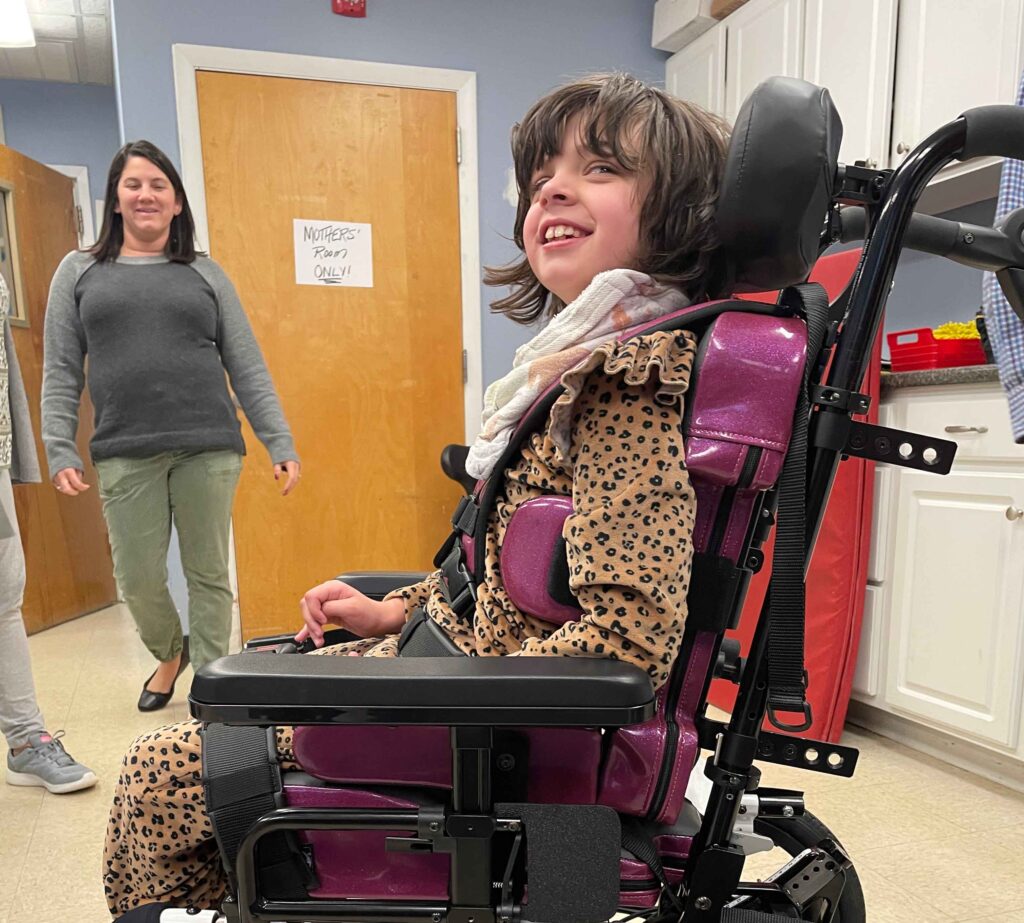 A young girl with shoulder-length brown hair, wearing a leopard-print outfit, smiles while sitting in a pink wheelchair. An adult woman with long dark hair is walking towards her in the background, in a room with white cabinets and a "Nurses' Pantry Only!" sign on the door.