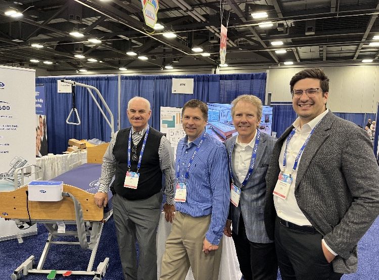 Four men, wearing business casual attire, stand together in an exhibition hall. Behind them is a booth displaying a medical bed and other equipment. The exhibition area is well-lit with various booths and banners in the background.