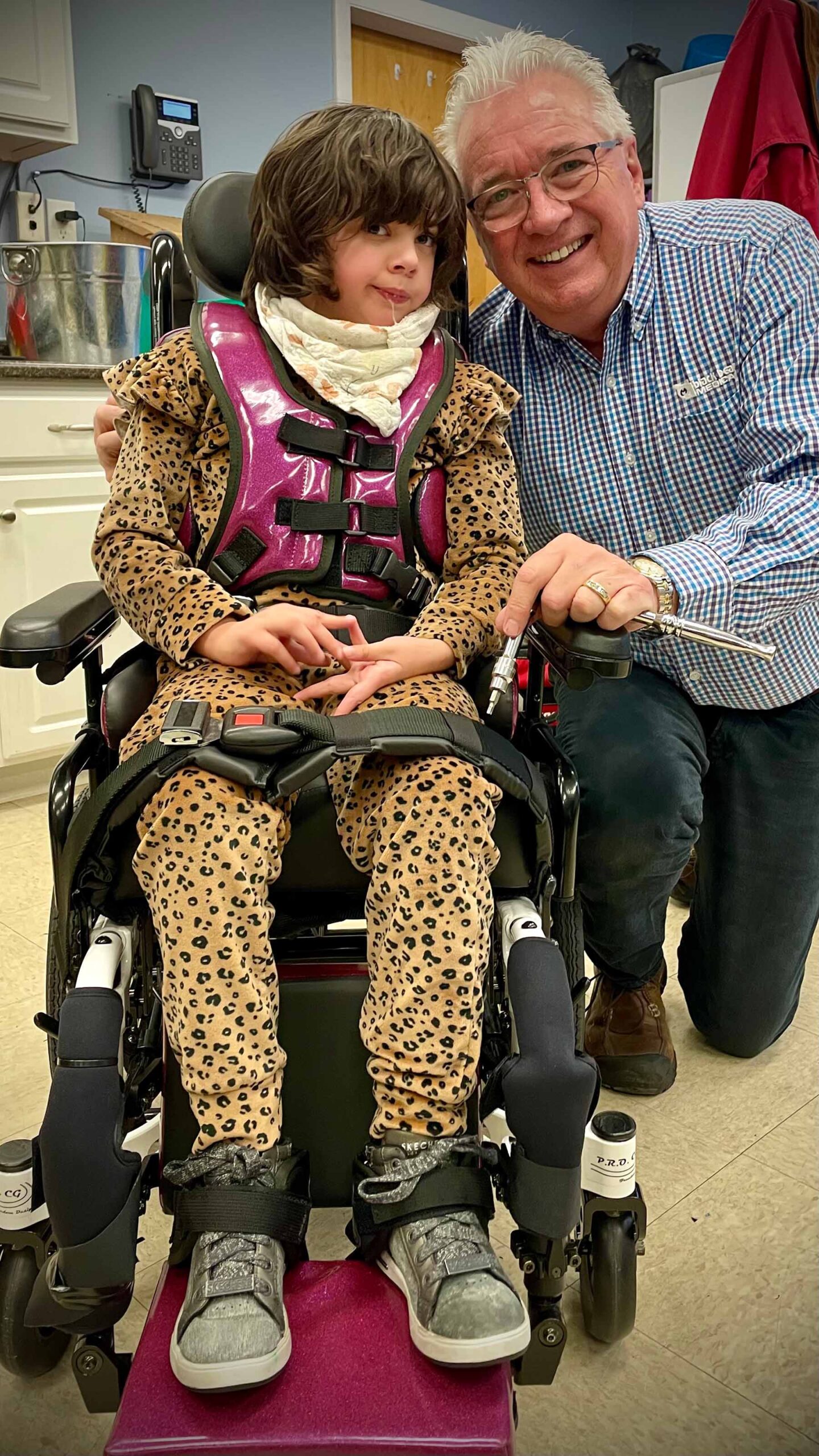 Aslynwearing a leopard-patterned outfit and a pink medical vest sits in a wheelchair, smiling and gesturing with their hands. An older person with white hair, wearing glasses and a checkered shirt, poses beside the child, holding one of their hands and smiling.