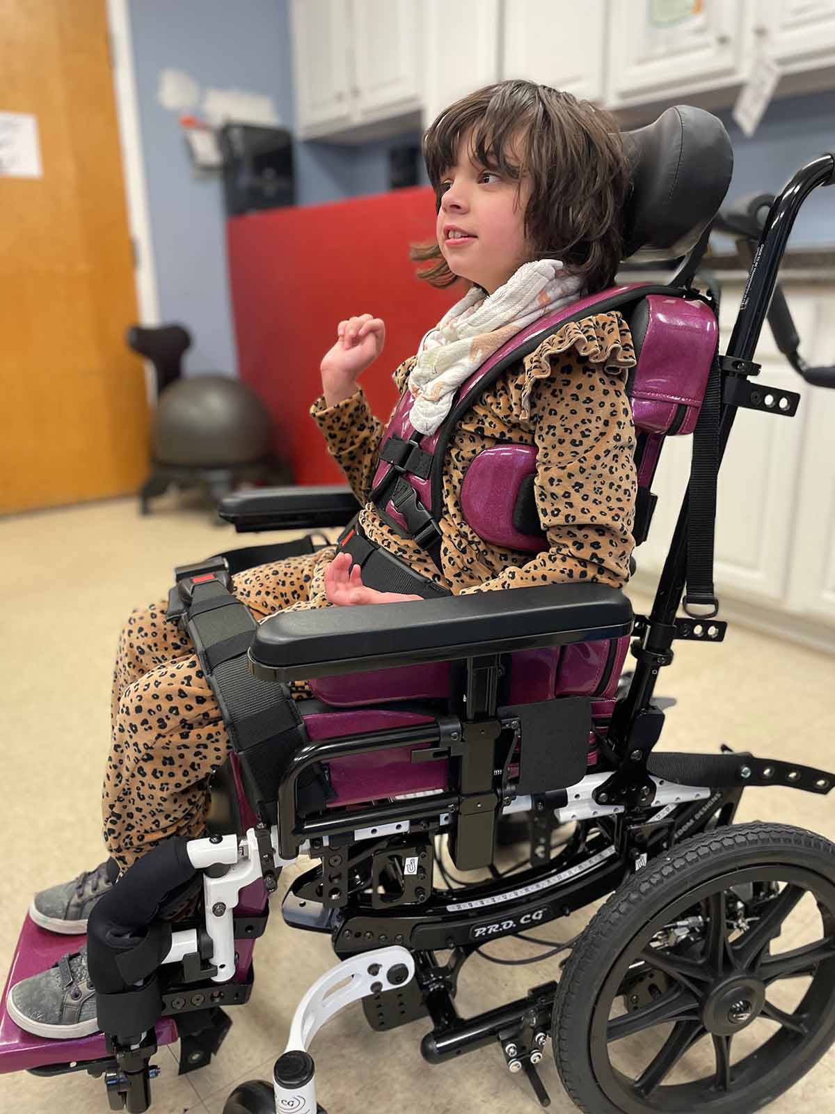 A young child with short brown hair sits in a specialized wheelchair. The child is wearing a brown leopard-print outfit and a white scarf. The room has light-colored walls, a wooden door, and various medical equipment. The child smiles slightly and appears engaged.