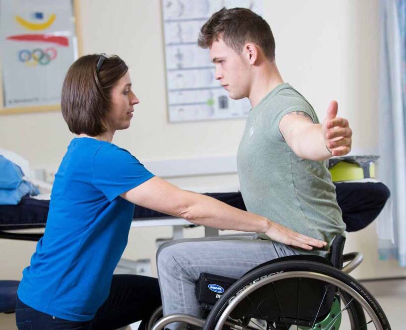 A physical therapist assists a man in a wheelchair by guiding his arm and supporting his back, helping him with arm mobility and posture exercises in a medical facility. Olympic rings and a medical chart are visible on the wall in the background.